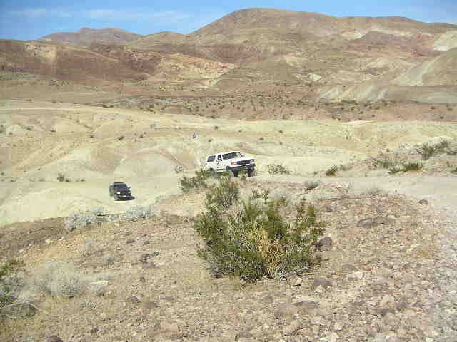White SUV and black truck on a dirt road in a desert with mountains in the background