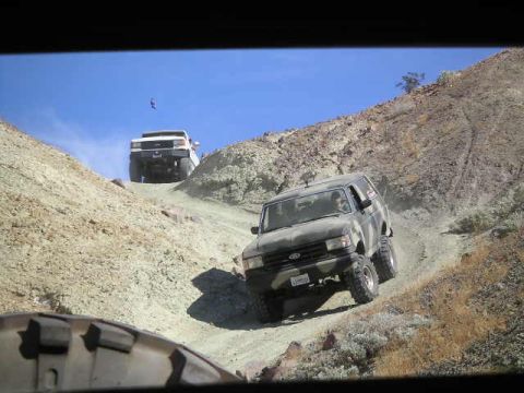 1988 Ford Bronco - A 1988 Ford Bronco navigates a steep, dusty trail in a mountainous desert landscape, with another white SUV visible further up the path.