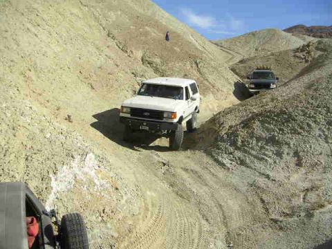 1988 Ford Bronco - Two SUVs navigate a steep, dusty trail through a desert canyon landscape.