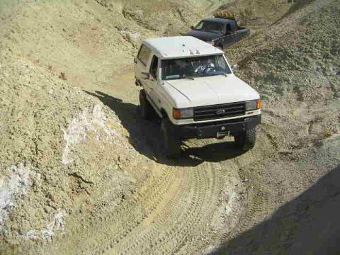 1988 Ford Bronco - Two trucks navigate a steep, sandy off-road trail in a desert-like environment.