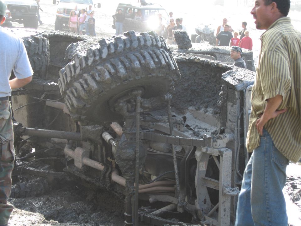 Overturned muddy off-road vehicle, front tire visible, with people watching