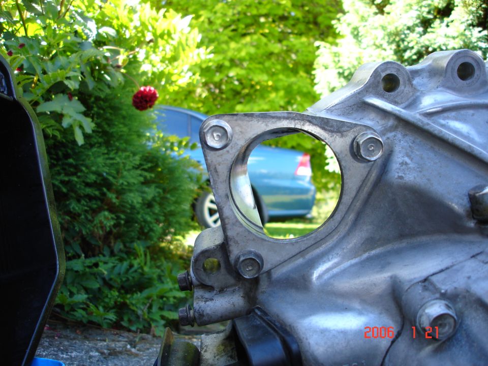 Aluminum engine casting with round open flange port; teal car visible through the hole in a garden setting