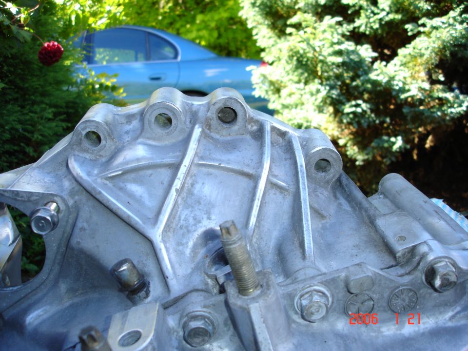 Bare aluminum transmission case close-up outdoors with blue sedan and greenery in background