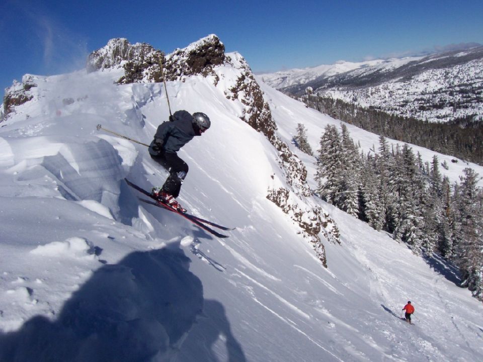 Skier jumping off a snowy cornice below a rocky ridge with mountains behind