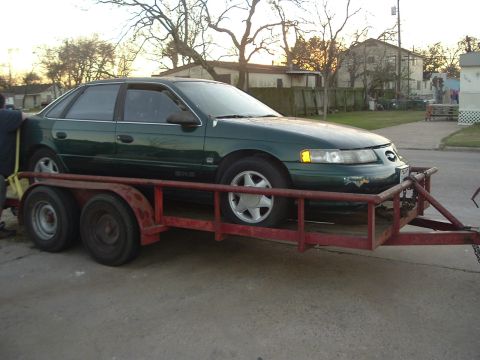 1993 Ford Taurus - A green sedan is being transported on a red trailer. The car's body style suggests a mid-1990s model.