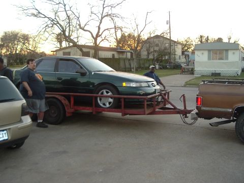 1993 Ford Taurus - A green sedan is loaded onto a trailer, hitched to a brown pickup truck, with two men observing.