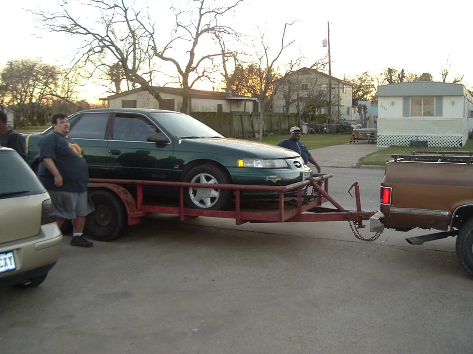 Green sedan on a trailer being towed by a brown pickup truck. Two men are standing nearby