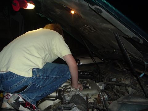 1993 Ford Taurus - A person crouches down to work on the engine of a vehicle with the hood open, illuminated by a work light.