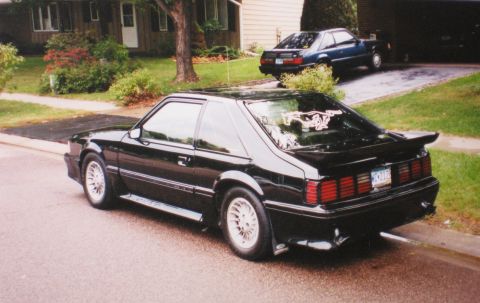 1990 Ford Mustang - A black Ford Mustang is parked on the street, with another blue car in the driveway behind it.