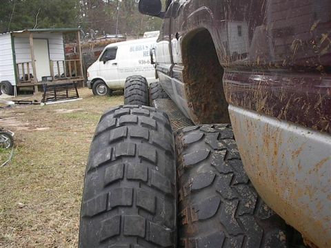 2001 Dodge 1500 - A maroon truck with large tires is parked next to a white van in a yard.