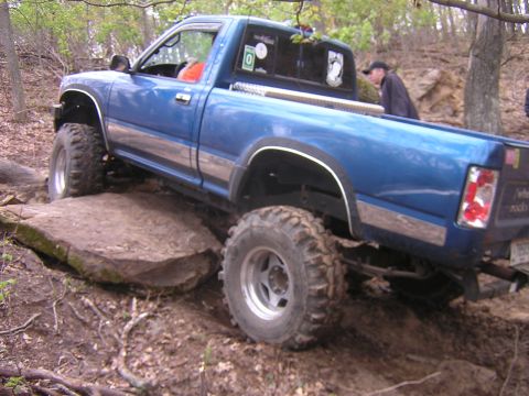 1977 Ford F-150 - A blue pickup truck is shown off-roading over large rocks and dirt.