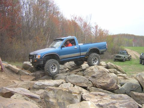 1977 Ford F-150 - A blue pickup truck is shown off-roading over a challenging rock obstacle with other vehicles in the background.
