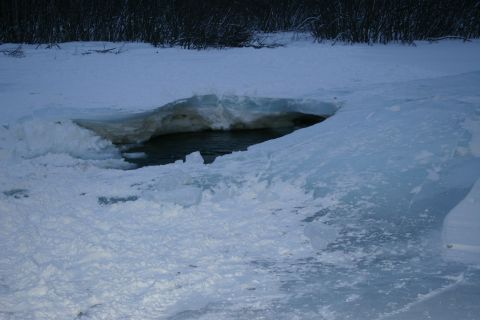 2003 Jeep TJ - A hole in the ice reveals dark water beneath the frozen surface, with trees visible in the distance.