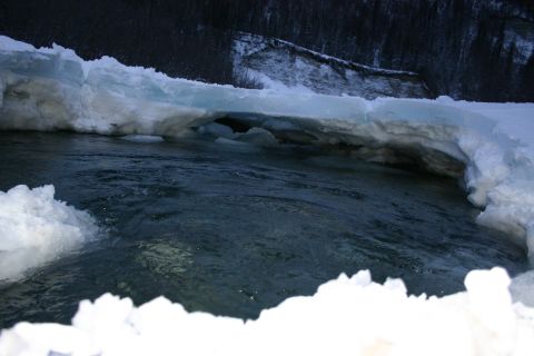 2003 Jeep TJ - A frozen river flows under ice formations, with snow-covered banks and dark trees visible in the background.