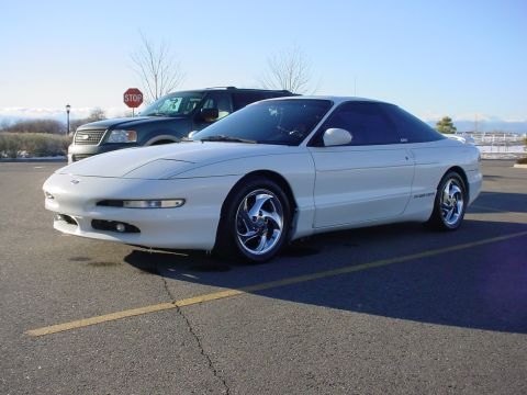 1996 Ford Probe - A white 1996 Ford Probe is parked in a lot with a dark SUV behind it under a clear sky.