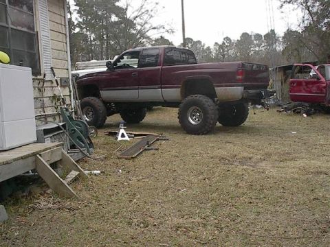 2001 Dodge 1500 - A lifted maroon 2001 Dodge 1500 pickup truck with oversized tires sits in a yard next to a house and another vehicle.