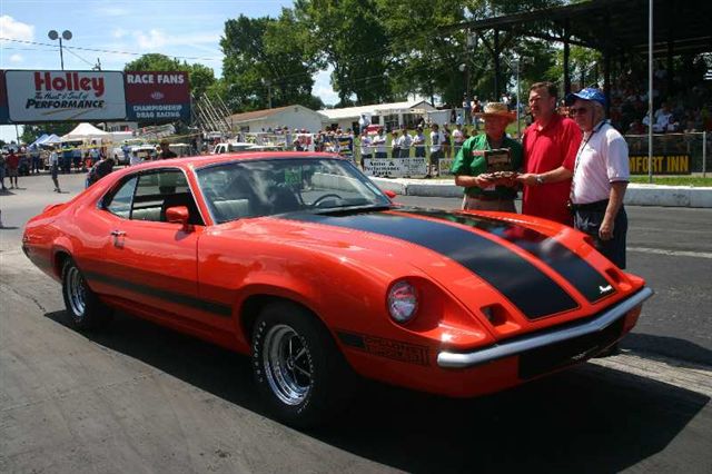 Orange fastback muscle coupe with black hood stripes at a drag race, men with trophy beside it