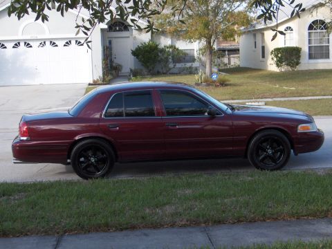 2004 Ford Crown Victoria - A maroon 2004 Ford Crown Victoria with tinted windows and black aftermarket wheels is parked curbside in front of a suburban house.