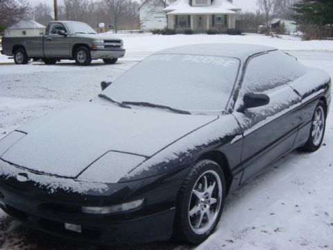 1995 Ford Probe - A dark Ford Probe sits covered in snow, with a pickup truck in the background on a cloudy day.