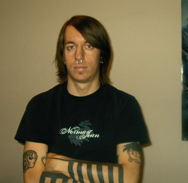 Man with nose ring, ear piercings, and extensive tattoos on arms and torso. He wears a black t-shirt with 'Norma Jean' printed on it