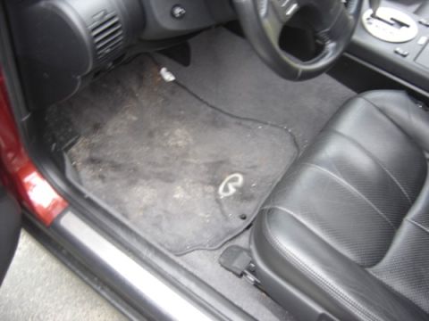 1999 Ford Taurus - The interior of a car shows a dirty floor mat with an Infiniti logo and a portion of the driver's seat.