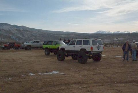 1986 Ford Bronco - Multiple modified SUVs and trucks are gathered in an open, dusty area with snow-capped mountains visible in the distance.