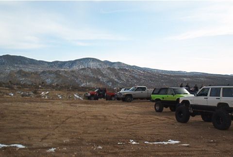 1986 Ford Bronco - Multiple modified trucks and SUVs are gathered on a dirt lot with snowy mountains visible behind them.