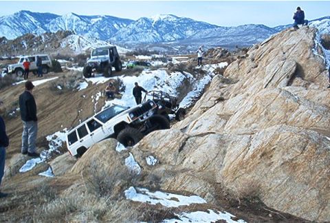 1986 Ford Bronco - Off-road enthusiasts tackle challenging terrain with snow-dusted rocks and mountains under a clear sky.