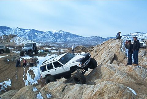 1986 Ford Bronco - Several off-road vehicles navigate a snowy, rocky terrain with mountains in the background.