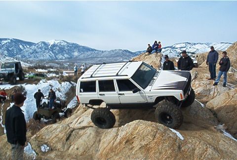 1986 Ford Bronco - A white Jeep Cherokee XJ navigates a challenging rocky trail with spectators observing the off-road action.