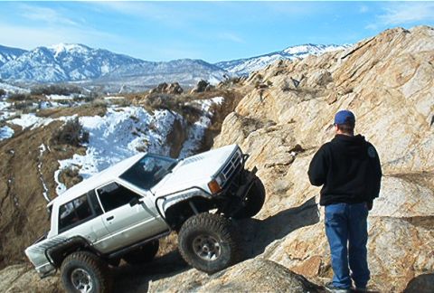 1986 Ford Bronco - A white SUV navigates a steep, rocky incline with snow-capped mountains visible in the distance.