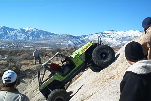 1986 Ford Bronco - An off-road vehicle navigates a steep, rocky terrain under a clear blue sky with snowy mountains in the distance.