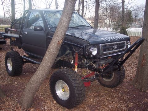 1995 Toyota Tacoma - Modified 1995 Toyota Tacoma pickup truck with custom off-road features and a flatbed, partially obscured by a tree.