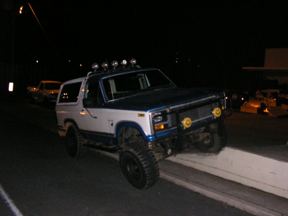 White and blue Ford Bronco with off-road tires and roof lights, angled on a curb at night