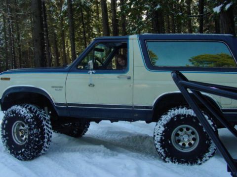 1986 Ford Bronco - A cream and blue Ford Bronco with large off-road tires is parked in a snowy forest.