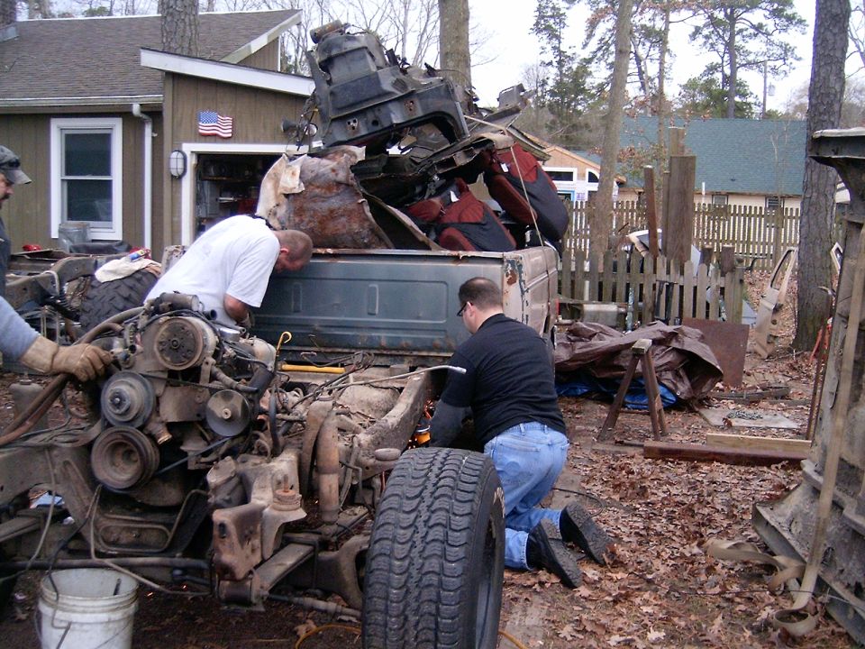 Two men dismantle a rusty pickup truck loaded with scrap parts in a backyard, sparks from a cutting torch visible.