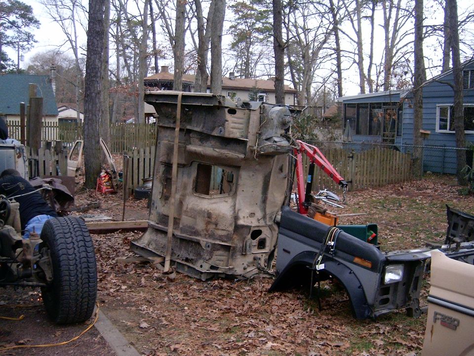 Disassembled truck: bare cab shell on its side, gray F250 front clip, and rolling frame in a backyard.
