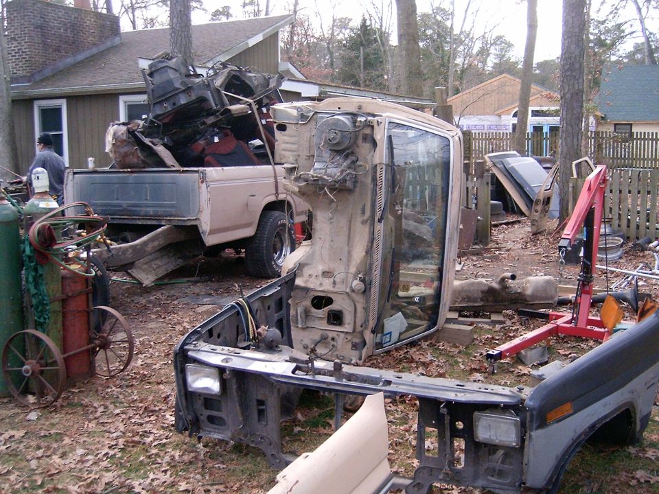 Disassembled tan truck cab and front clip in a backyard, with a parts-loaded pickup and engine hoist behind