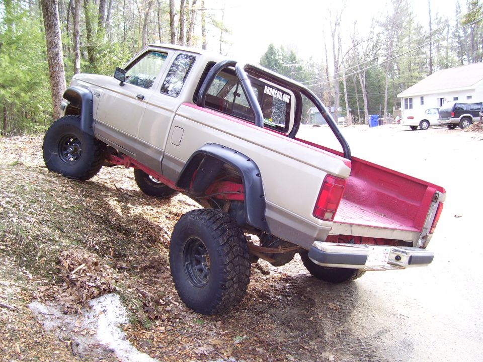 Tan lifted Ford Ranger pickup with roll bar, rear three-quarter on leaf-covered driveway