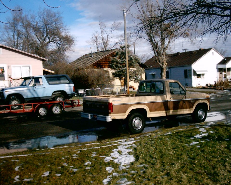 Light blue Ford Bronco on a flatbed trailer behind a tan two-tone Ford pickup on a residential street