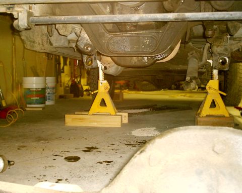 1985 Ford Bronco - Vehicle's undercarriage is supported by jack stands in a garage setting, revealing mechanical parts.