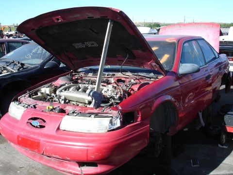 1993 Ford Taurus - A red Ford Taurus SHO sits in a junkyard with its hood open, revealing the engine and other components.