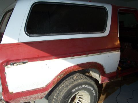 1978 Ford Bronco - Rear quarter of a two-tone red-and-white full-size Ford SUV in a garage, showing faded paint, a rust-eaten wheel arch lip and old body fill…