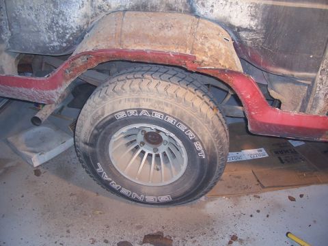 1978 Ford Bronco - Rear wheel well of a stripped truck body in a garage, with the quarter panel sanded to bare metal and rust above a faded red rocker and bod…