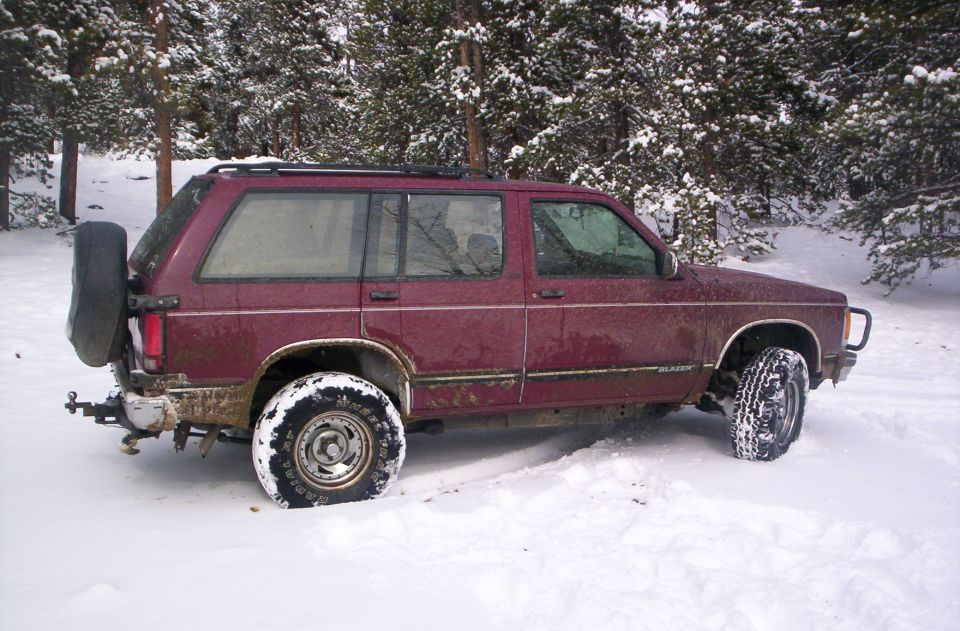 Maroon Chevrolet S10 Blazer SUV with white-letter tires parked in deep snow among pine trees