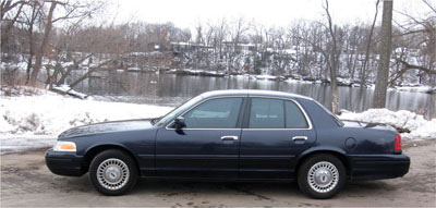 1999 Ford Crown Victoria - A dark blue Ford Crown Victoria is parked on a dirt road next to a river during winter.