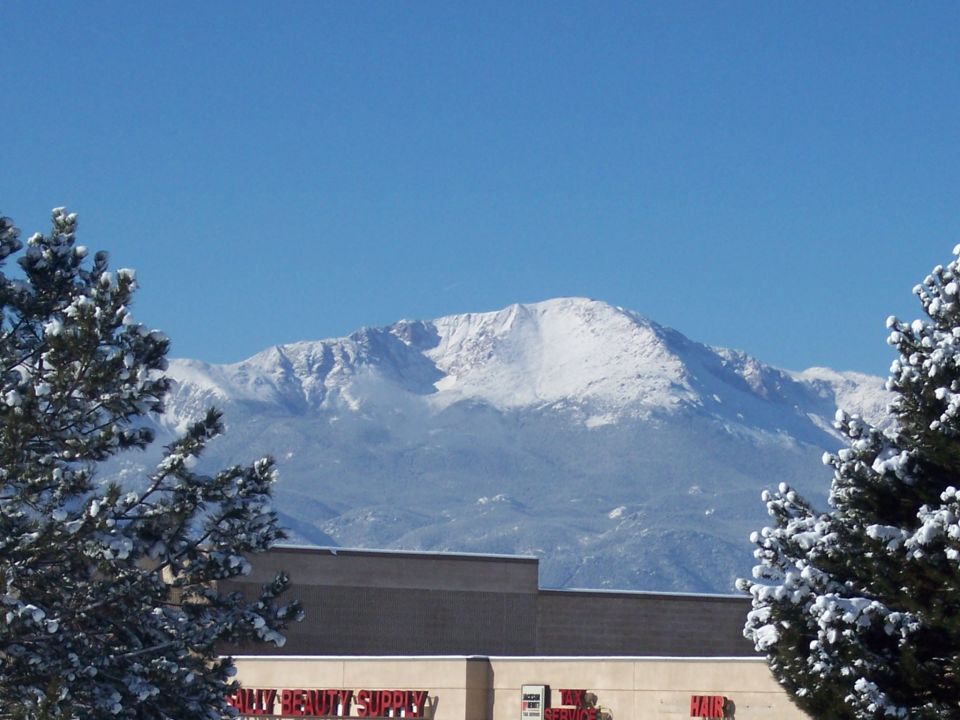 Snow-capped mountains under a clear blue sky, with pine trees and a commercial building in the foreground. Storefront signs include 'SALLY BEAUTY SUPPLY'