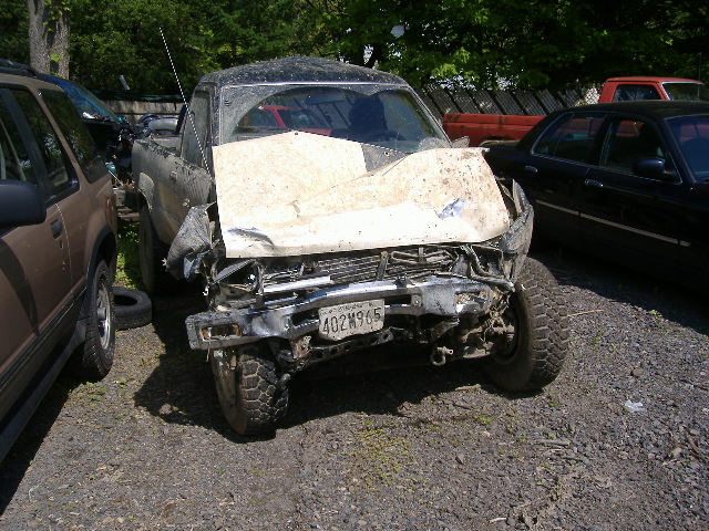 Front view of a heavily damaged pickup truck with a bent hood and grille. License plate reads 402M965. Another vehicle is parked to the left