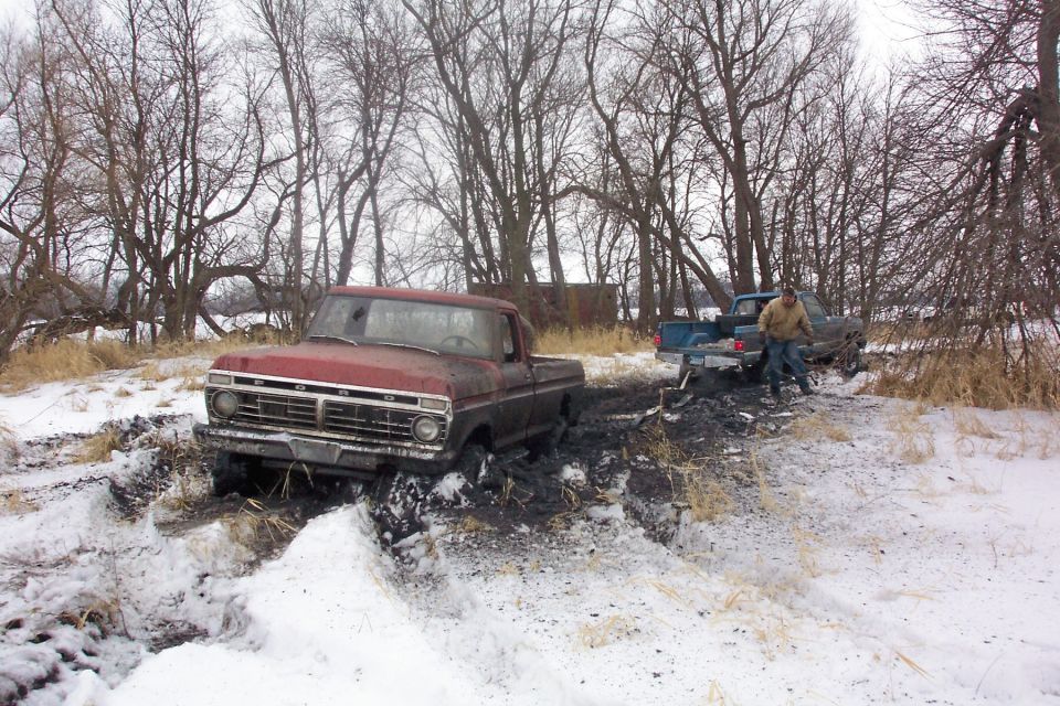 Red pickup truck stuck in mud and snow, another truck behind it with a person standing nearby