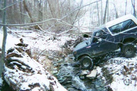 1986 Ford Bronco - A Ford Bronco traverses a rocky, snow-covered stream in a wooded area.